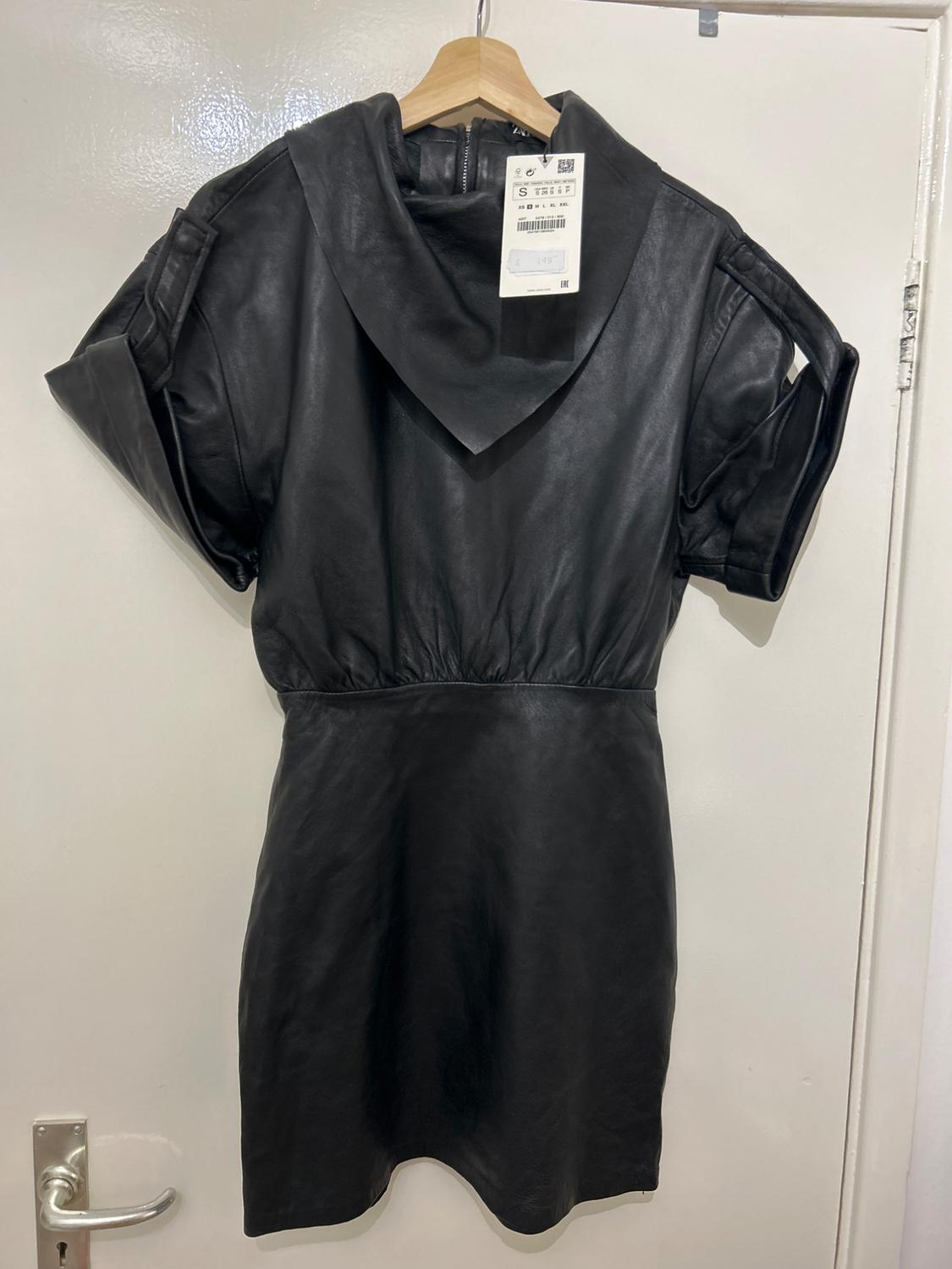 Black leather dress with a hood and short sleeves, hanging on a wooden hanger. Visible tag with product details.