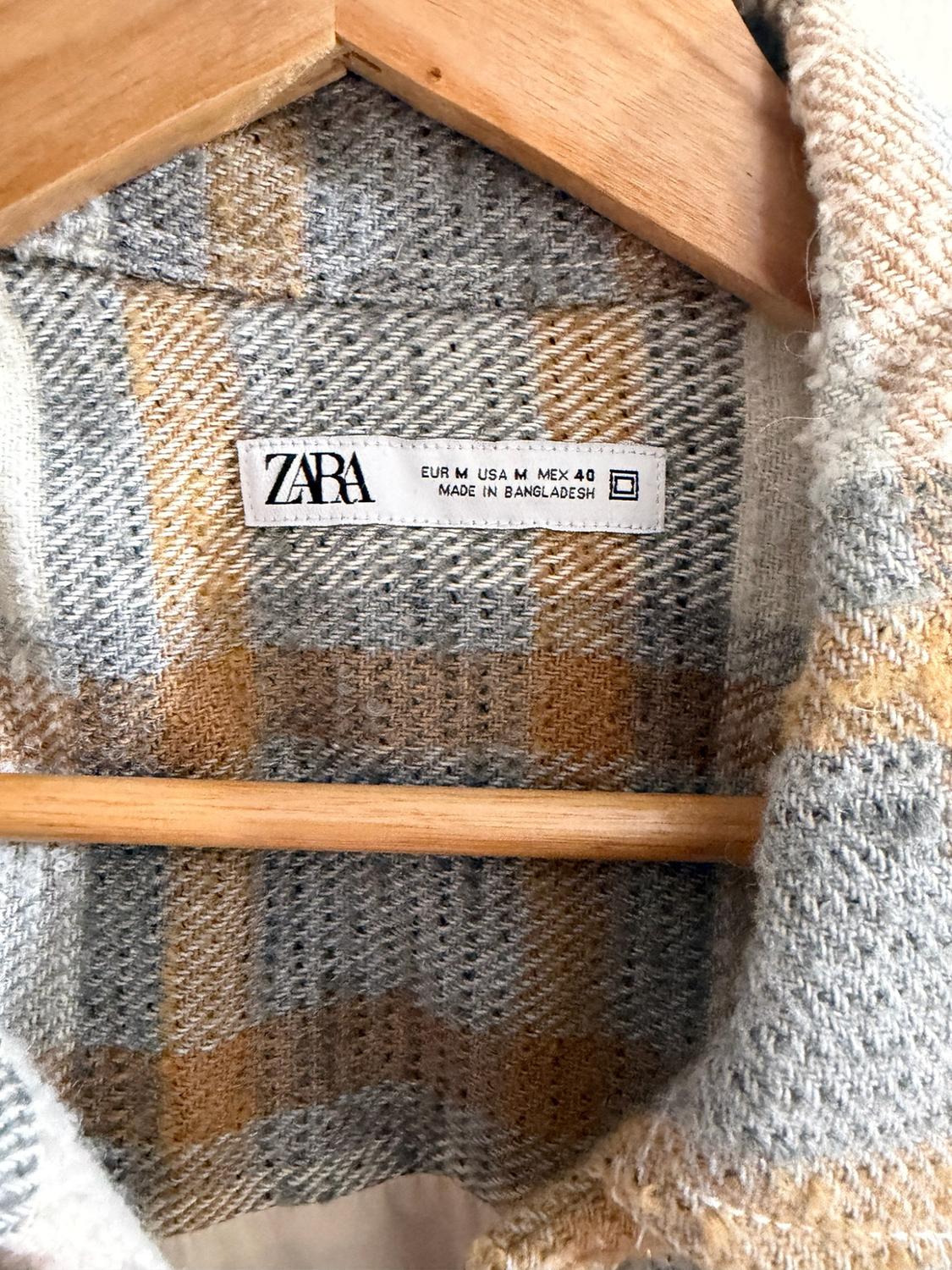 Detail of grey and mustard check garment on a wooden hanger. Text: ZARA EUR M USA M MEX 40 MADE IN BANGLADESH.