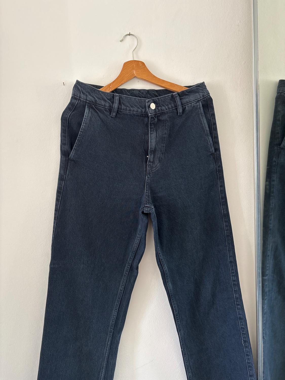 Dark blue jeans hanging on a wooden hanger against a white wall. To the right, the reflection of the garment can be seen in a mirror.