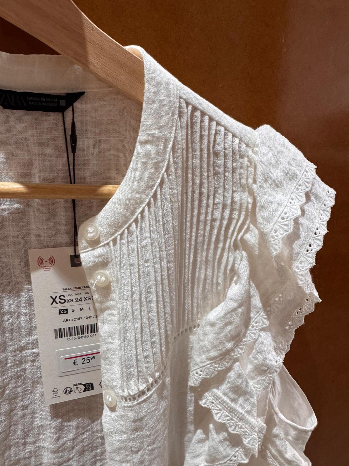 White linen blouse with pleated details and lace on the sleeves, hanging on a wooden hanger. Tag shows price of €25.95.
