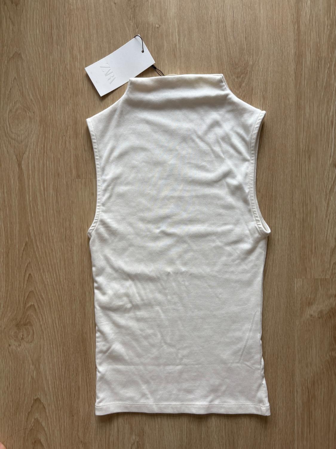 White sleeveless high-neck top on a wooden floor. Label with the text ZARA.