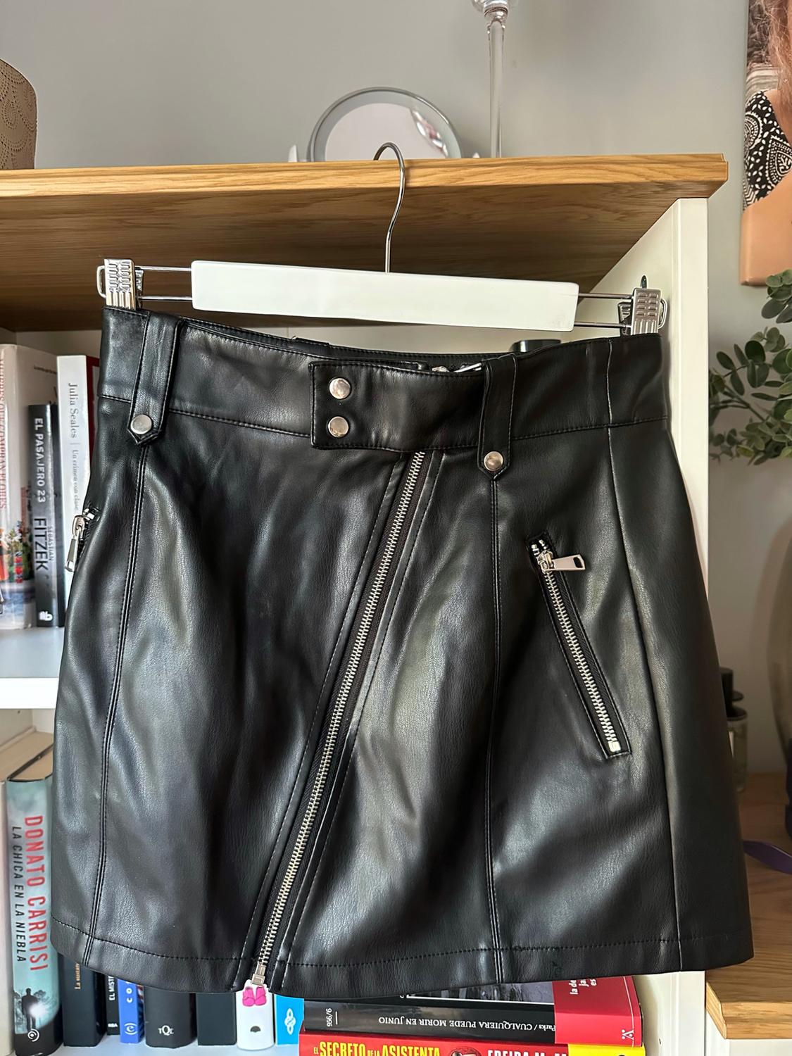 Black leather-effect skirt with an asymmetric zip hanging on a bookcase with books: THE PASSENGER 23 FITZEK, THE HOUSEMAID’S SECRET.