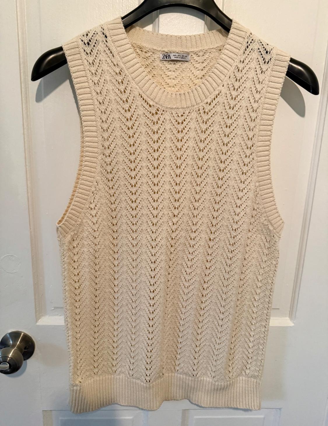 Beige knitted sleeveless waistcoat with a herringbone design, hanging on a black hanger over a white door. Label visible.