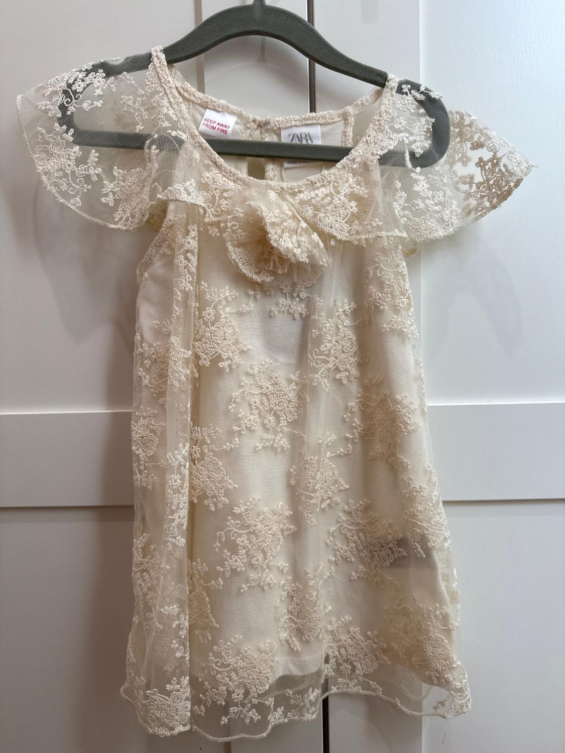 Beige lace dress hanging on a grey hanger. Visible label with text: "KEEP AWAY FROM FIRE" and "ZARA". White background.