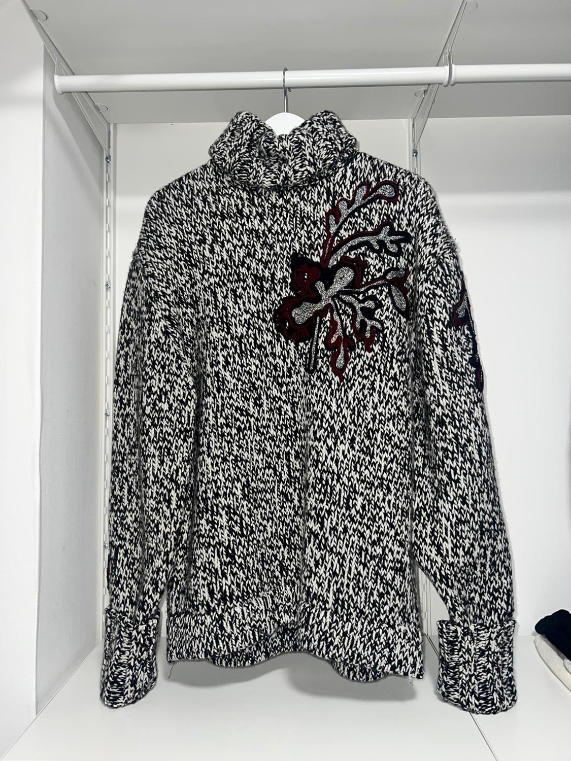 Black and white knitted polo neck jumper with a red and black floral design on the chest, hanging in a white wardrobe.