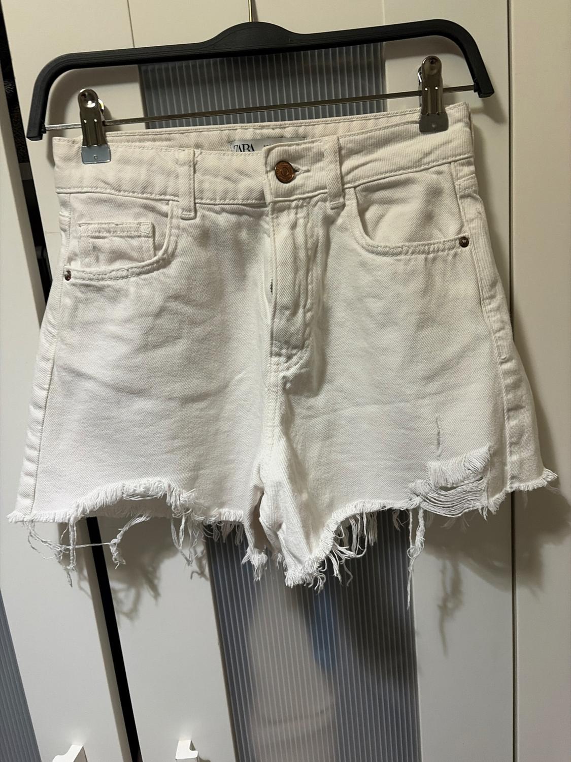 White denim shorts with a frayed hem, hanging on a black hanger in front of a white wardrobe.
