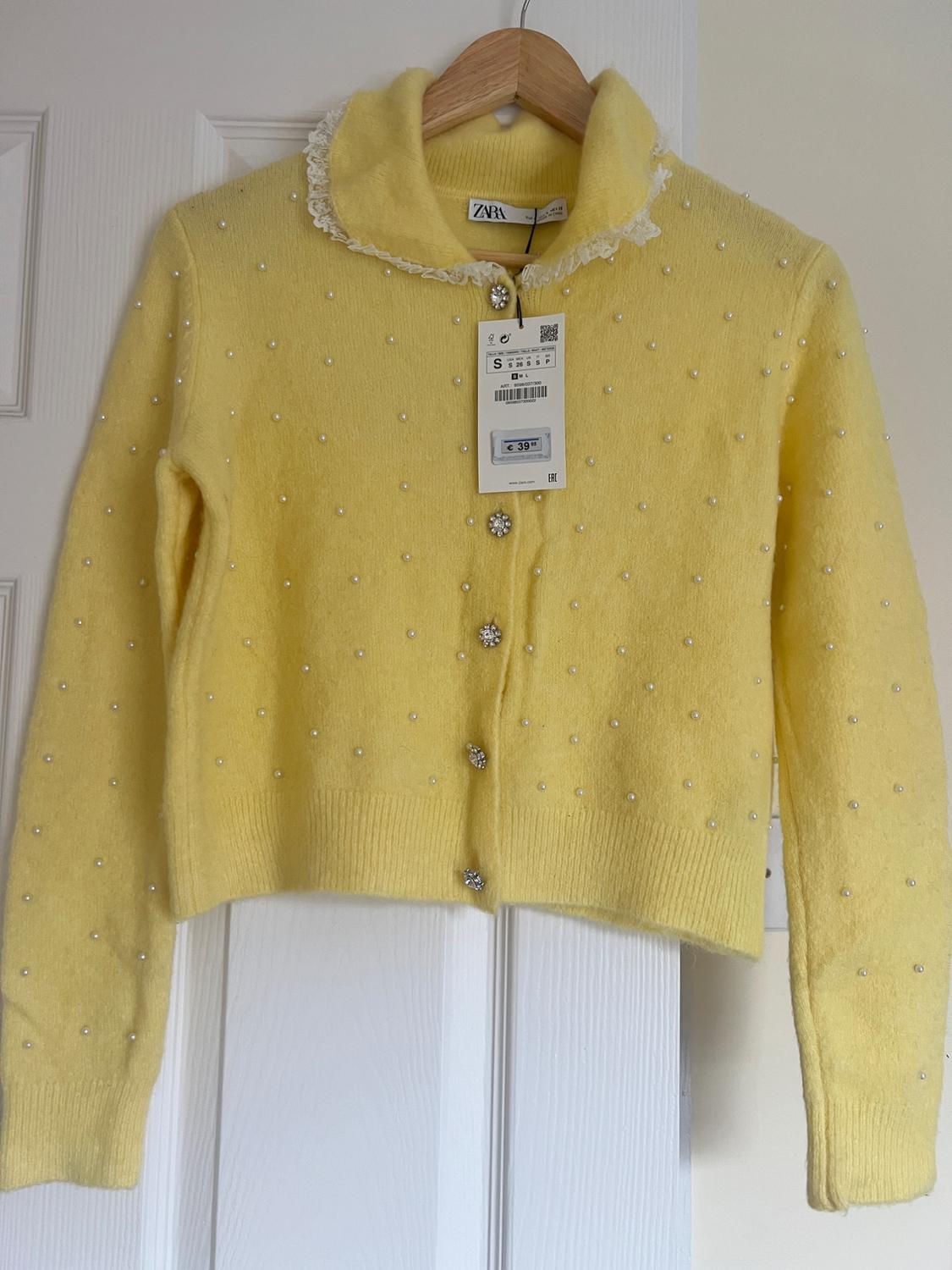 Yellow cardigan with pearls and decorative buttons, hanging on a hanger. Visible tag with price of €39.95.