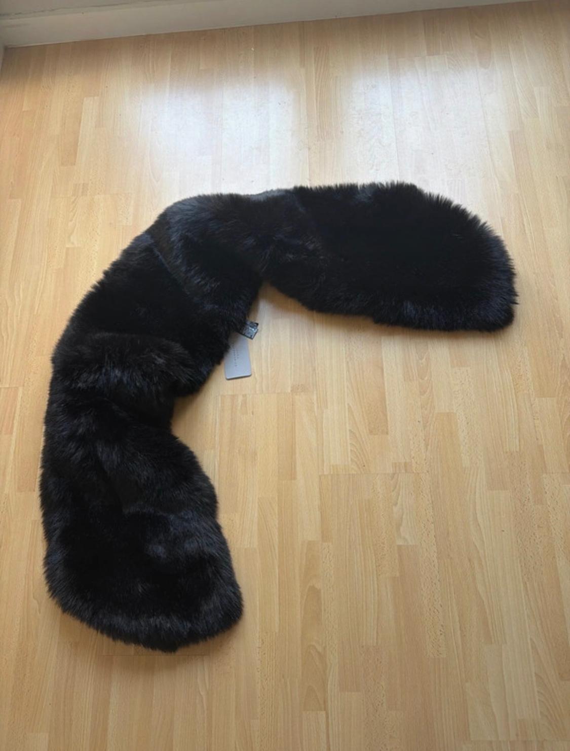 Black faux fur scarf on light wooden flooring. Visible tag attached to scarf.
