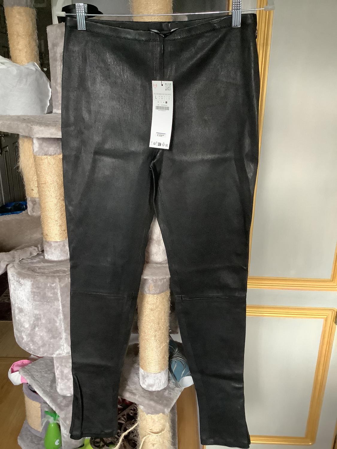 Black trousers hanging on a clothes rail, with a visible label in the centre. Cat scratching post and plain wall in the background.