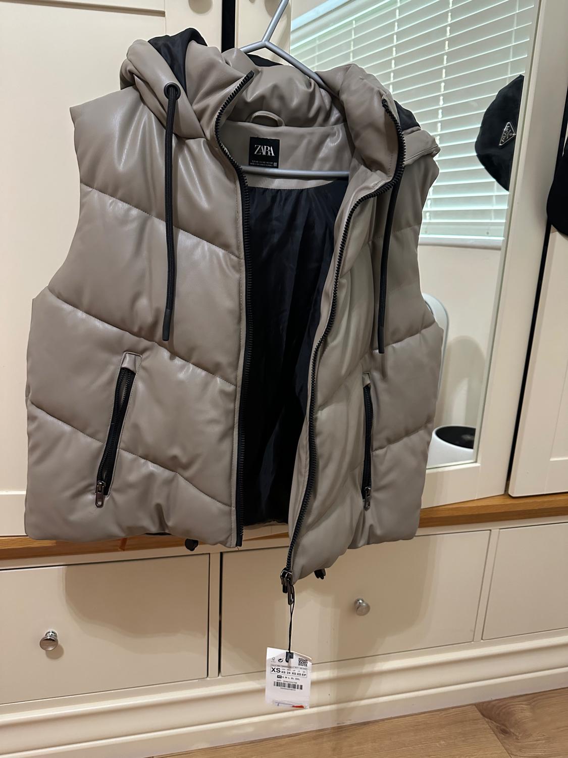 Grey padded gilet with hood and black zips, hanging on a hanger. Visible label with size XS. White wardrobe background.