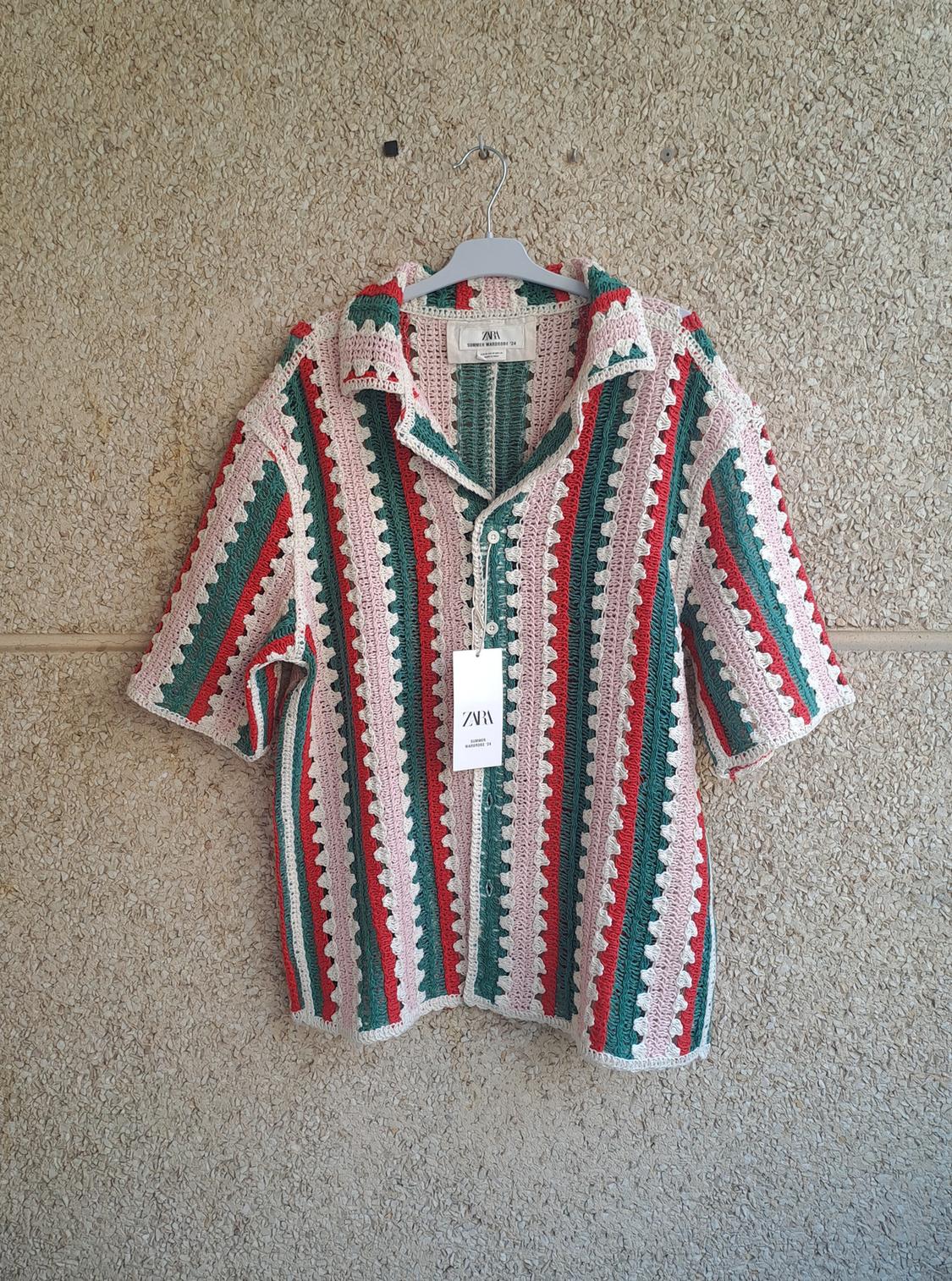 Crochet shirt with vertical red, green, and white stripes, hanging on a textured wall. Visible label with text "ZARA".