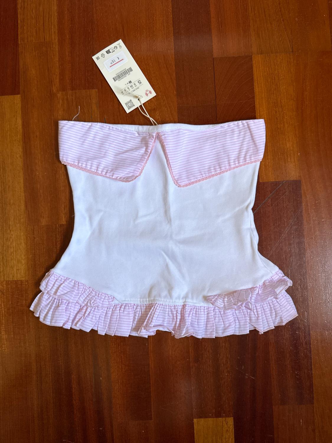 White strapless top with pink stripe detail on the neckline and ruffles on the hem, on a wooden floor.