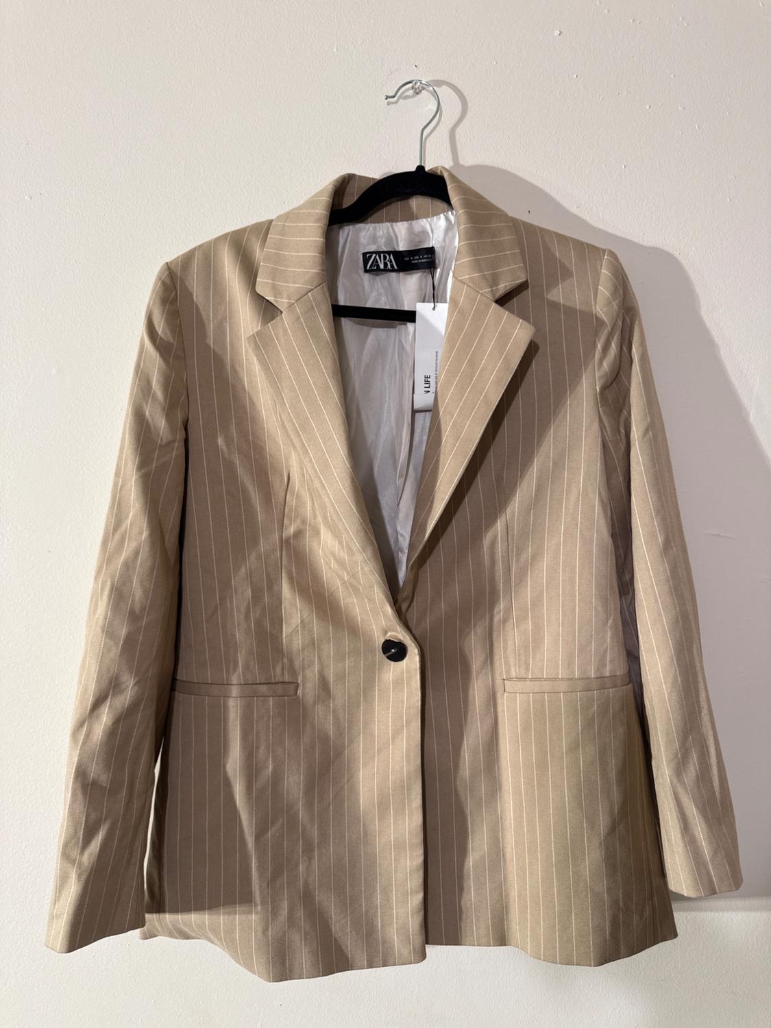 Beige jacket with vertical stripes hanging on a hanger. Visible label with the text "ZARA". White background.