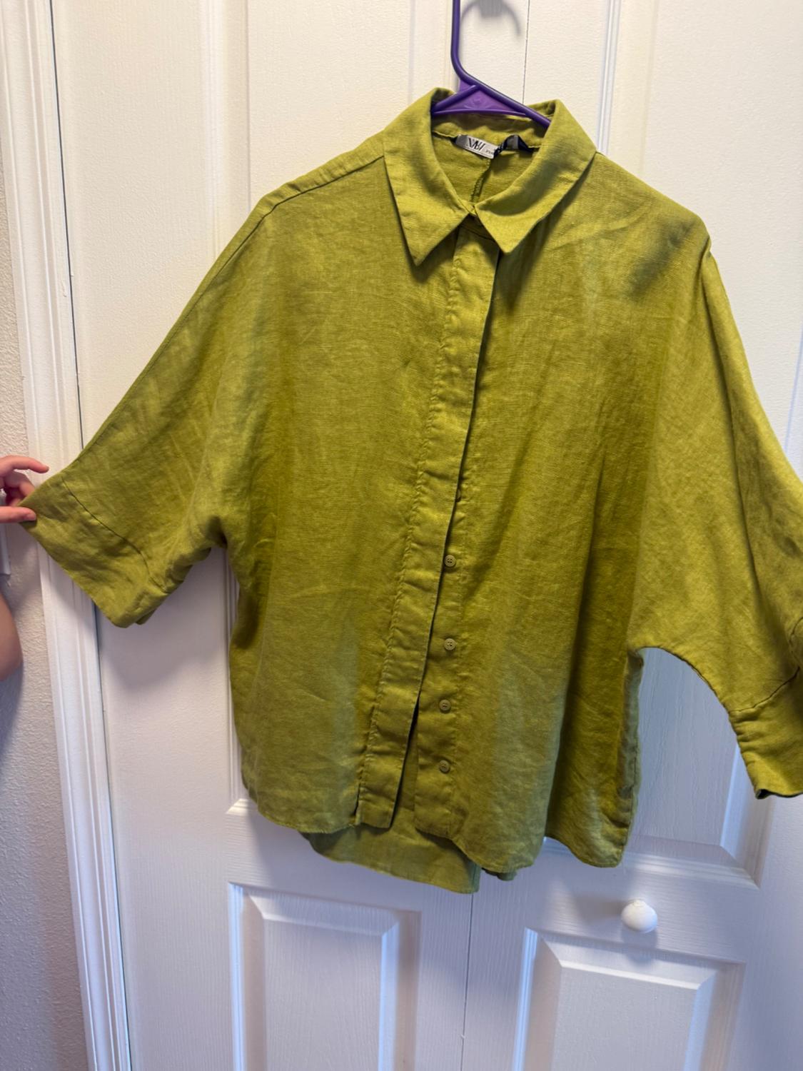 An olive green short-sleeve shirt is hanging on a purple hanger in front of a white door. A hand is holding the sleeve on the left side.