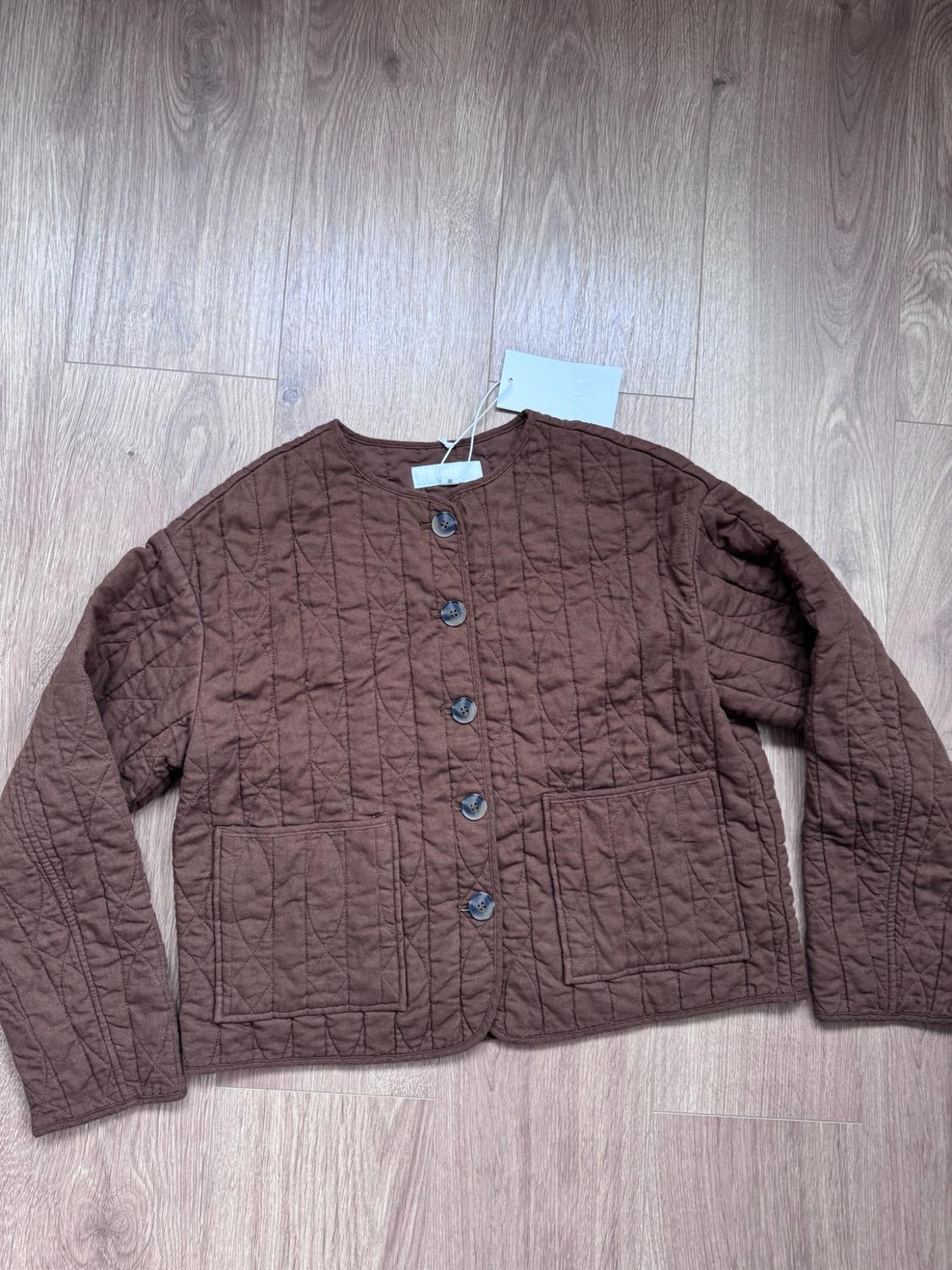 Brown quilted jacket with a round neck, button fastening and front pockets, laid out on a light wooden floor.