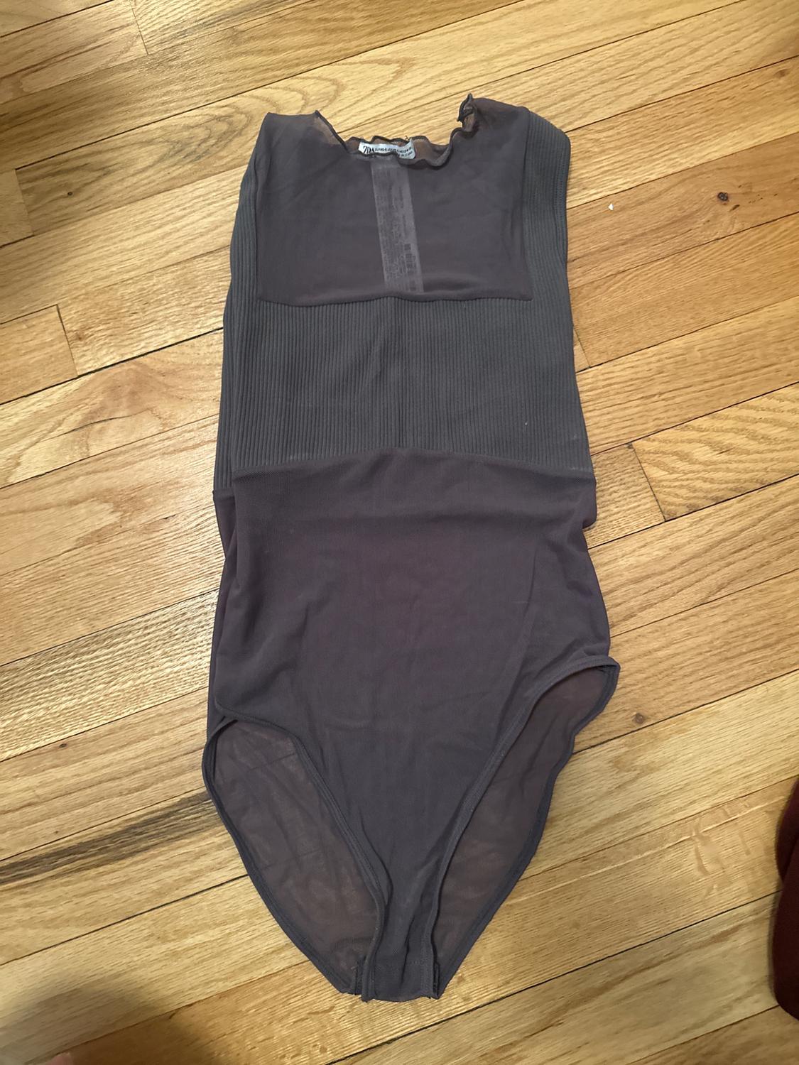 Sleeveless black bodysuit on a wooden floor. Simple design with ribbed texture on the chest and a round neck.