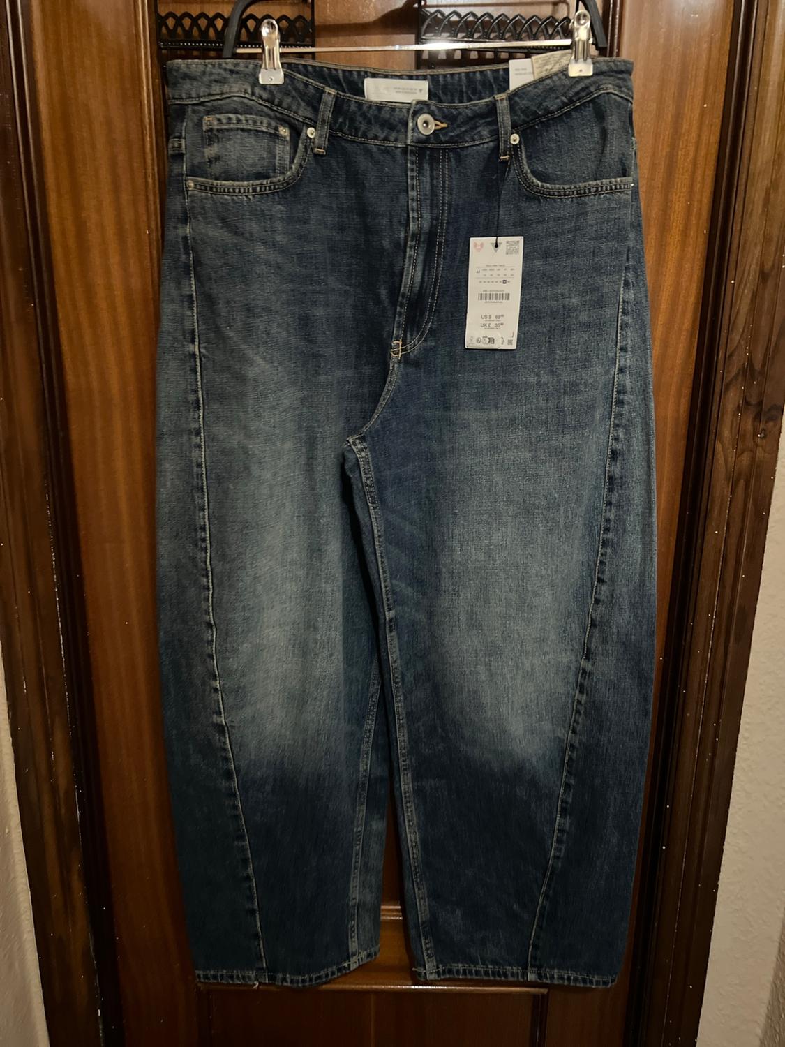 Wide blue denim trousers with front seams and a washed effect, hanging on a hanger in front of a wooden door with its tag.