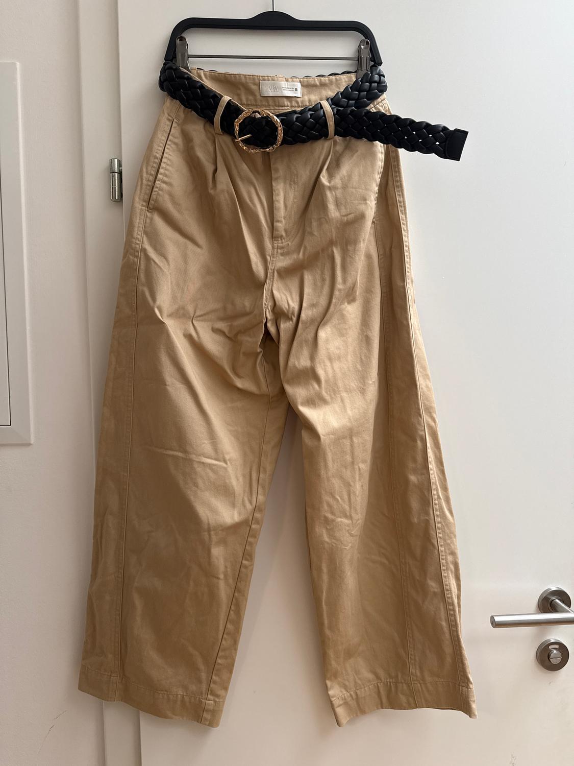 Beige trousers hanging on a white door, with a black braided belt and gold buckle.