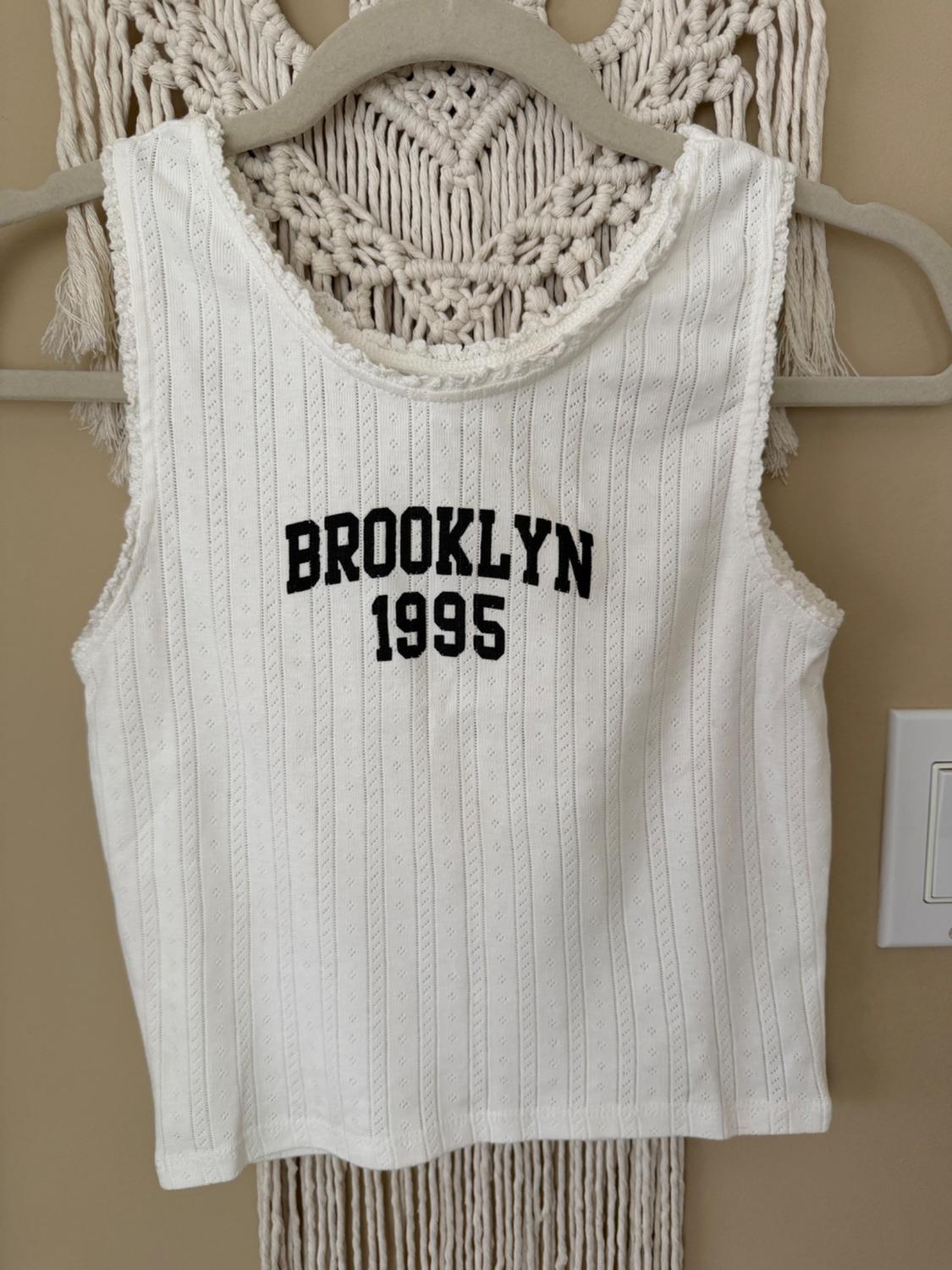 White sleeveless t-shirt with "BROOKLYN 1995" text in black, hanging on a hanger against a beige macrame background.