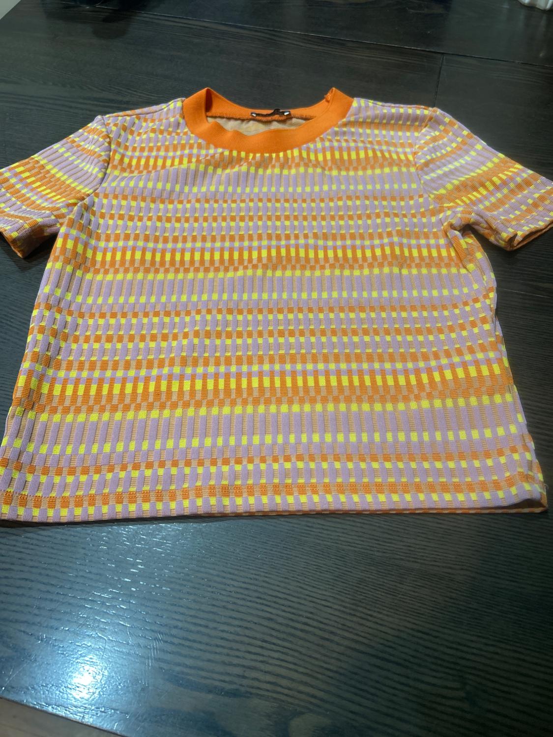 Short-sleeve t-shirt on dark table, with geometric print in shades of orange, yellow, and lilac, and solid orange collar.