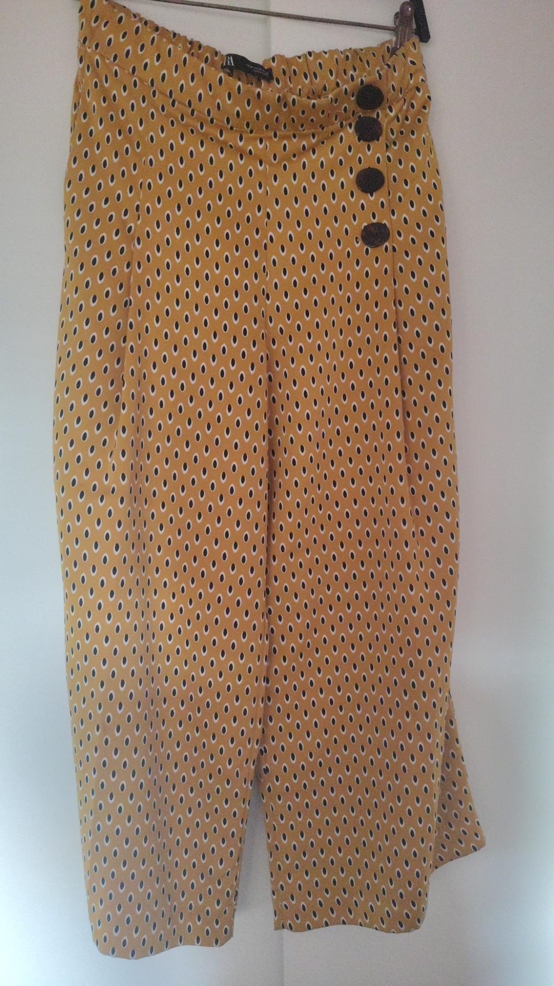 Yellow trousers with a geometric print, elasticated waist, and 4 side buttons, hanging on a hanger against a white background. Text: ZARA.