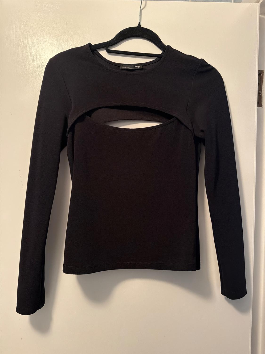 Black long-sleeved top with a front opening, hanging on a black hanger over a white door. Visible tag: ZARA.