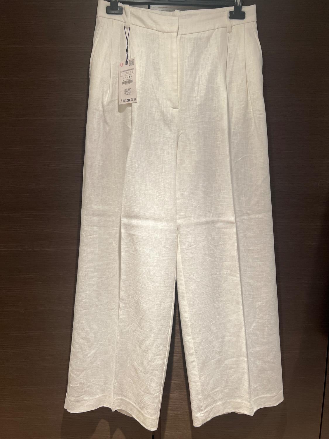 White wide-leg tailored trousers with a tag, hanging on a black hanger against a dark wood background.