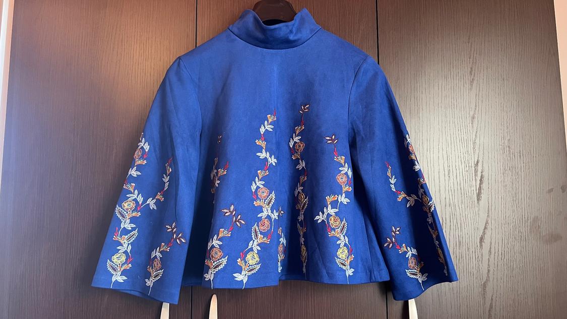 Blue jacket with colourful floral embroidery on the sleeves and front, hanging on a hanger in front of a dark wardrobe.