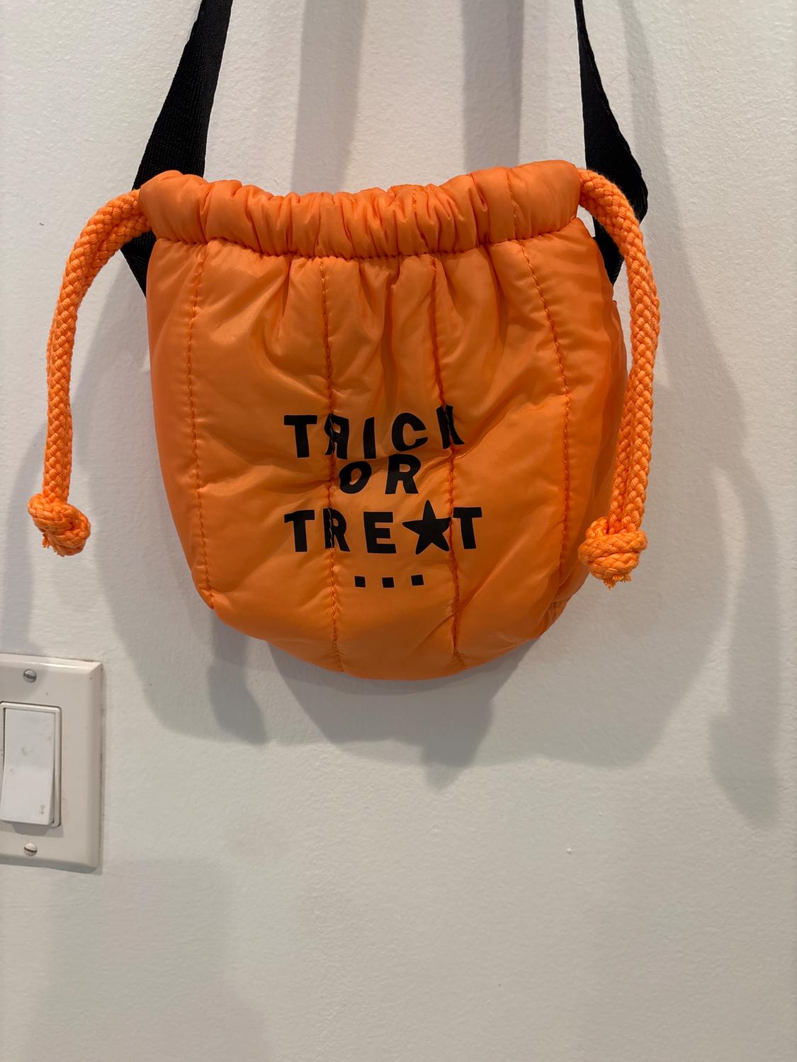 Orange bag with drawstrings and black "TRICK OR TREAT" text, hanging on a white wall next to a light switch.