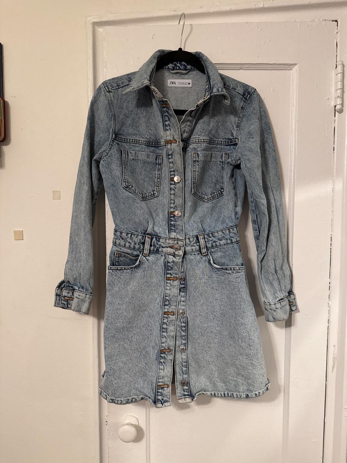 Light blue denim dress with front buttons and pockets, hanging on a white door.