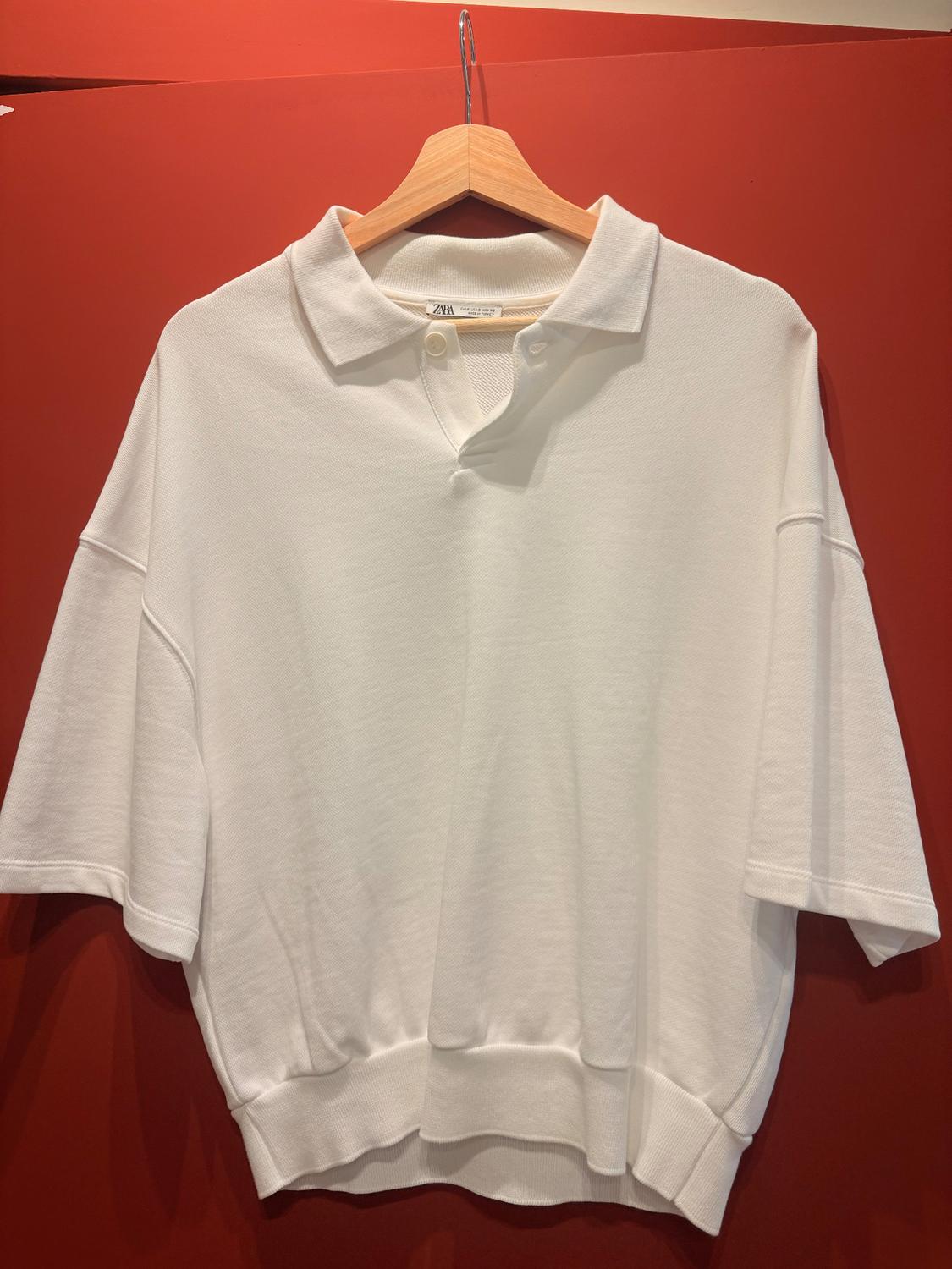 White short-sleeved polo shirt on a wooden hanger, hanging against a red background. Visible label on the collar.