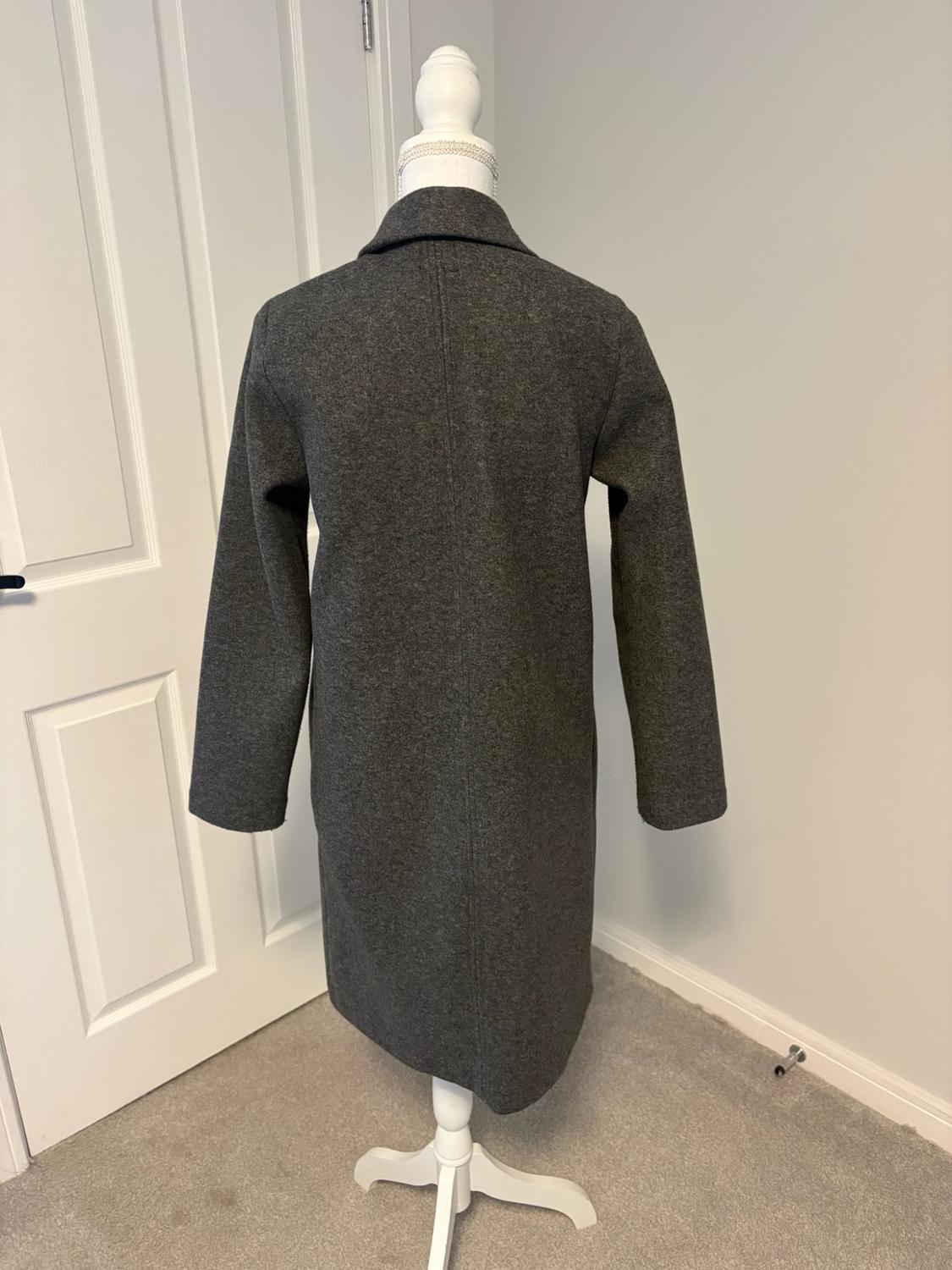 Rear view of a dark grey long coat on a white mannequin in a room with light-coloured walls, a white door, and a grey carpet.