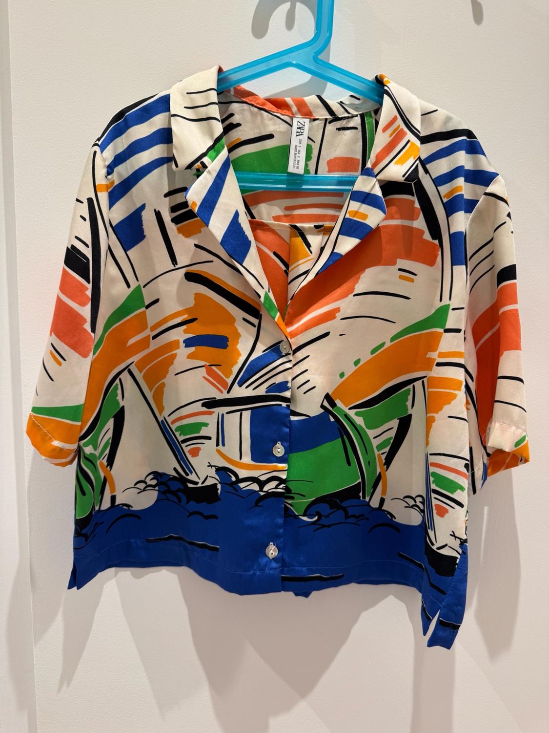 Short-sleeved shirt with an abstract print of boats and waves, hanging on a blue hanger against a white background. Text on the label: zara.