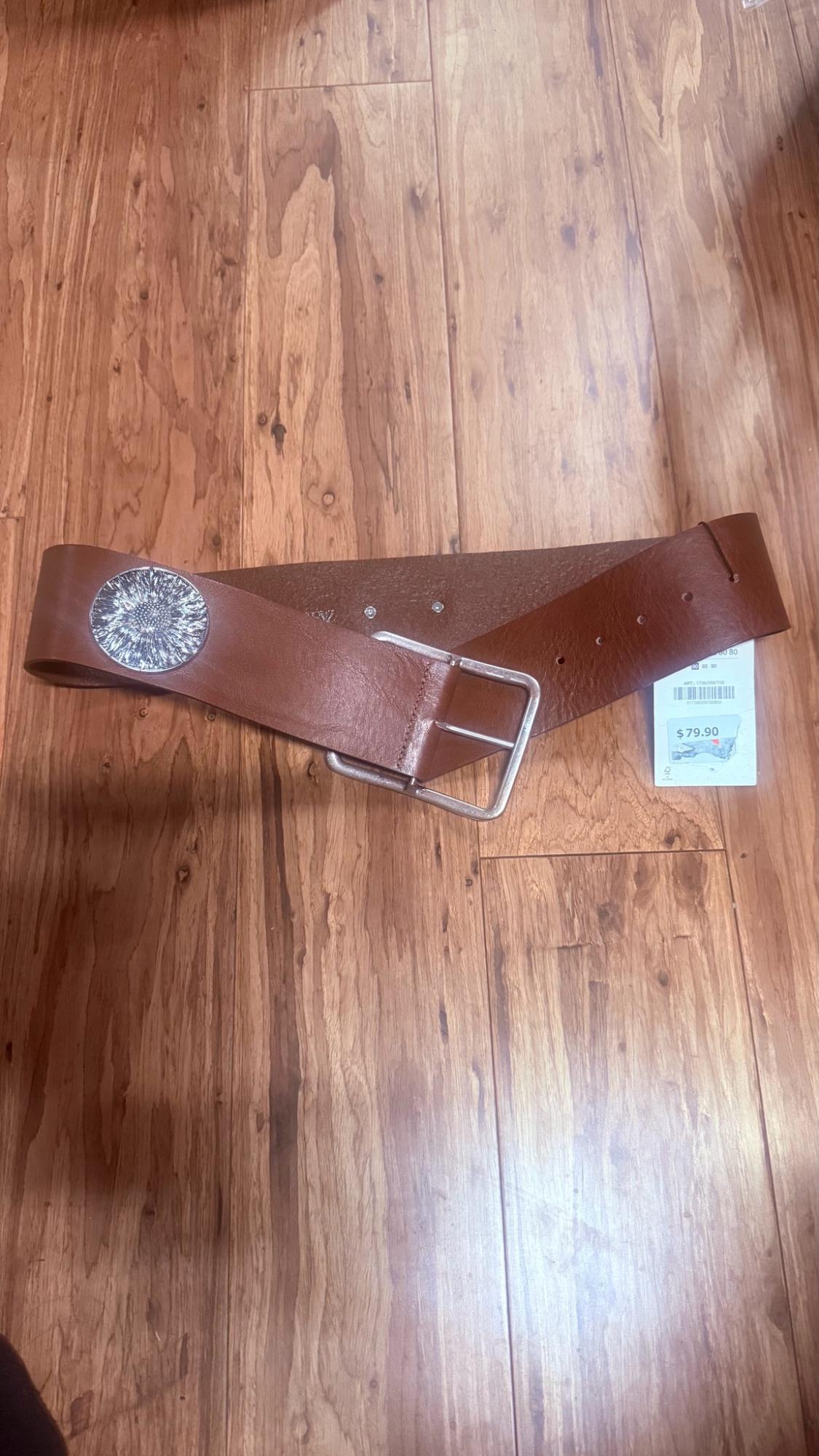 Wide brown belt with a square silver buckle and round embellishment on a wooden floor. Tag with the text "$79.90".