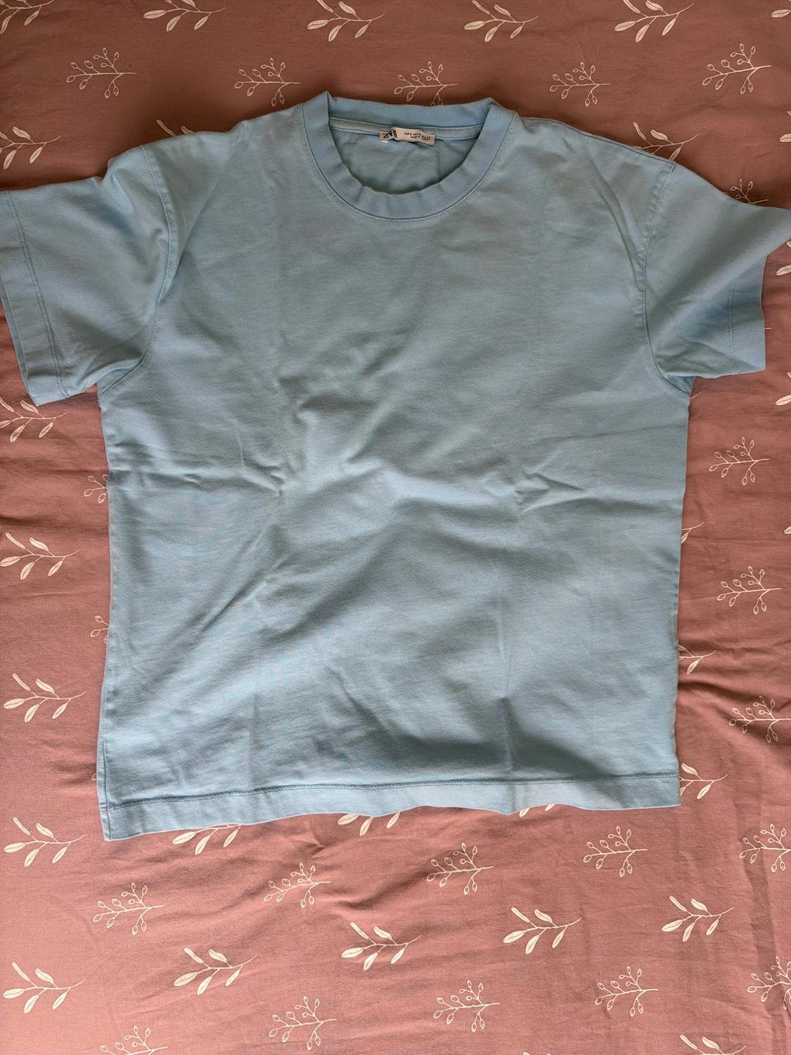 Light blue short-sleeve t-shirt on a pink fabric background with a white leaf print.