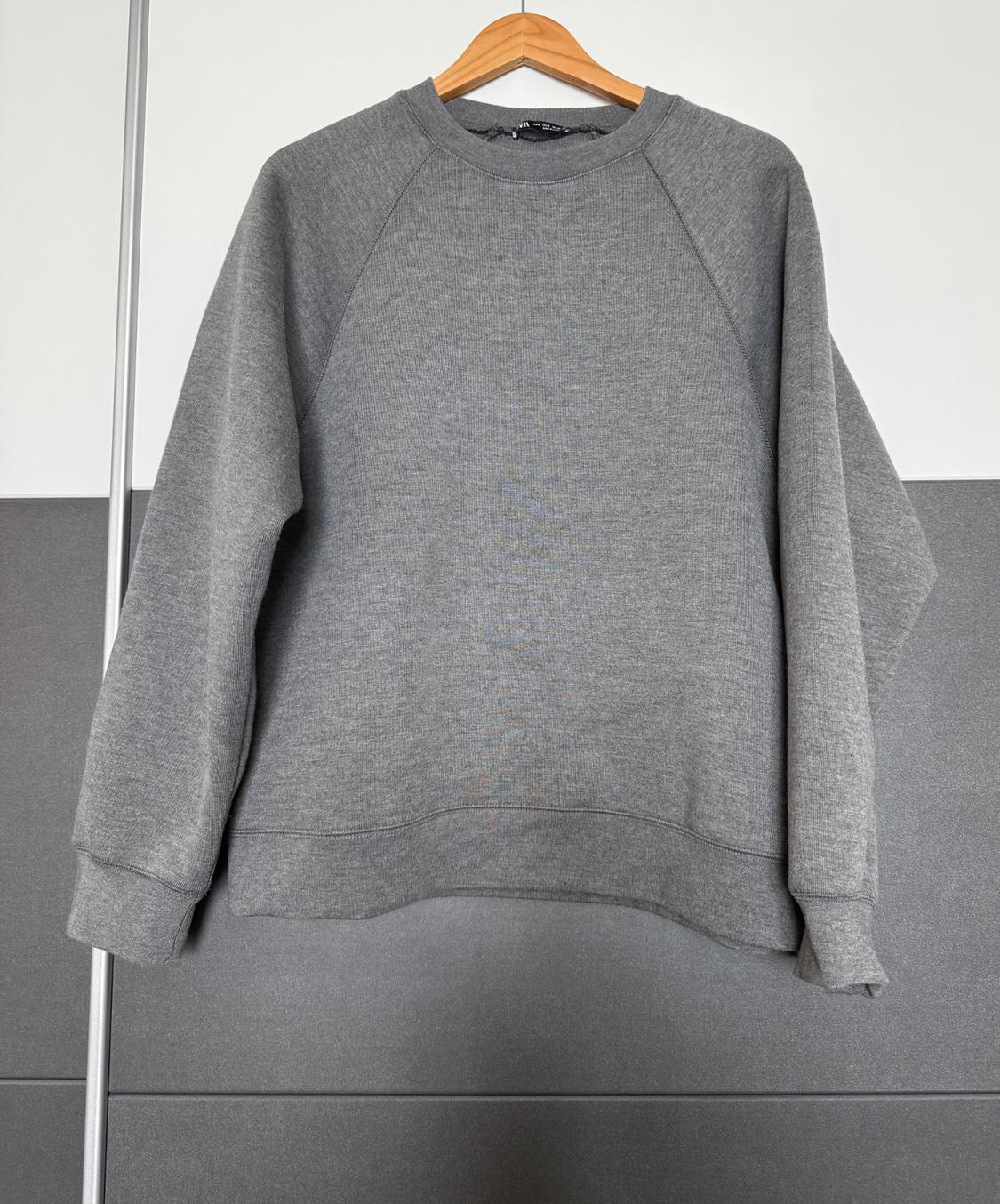 Grey long-sleeved sweatshirt hanging on a wooden hanger, grey and white wall background.