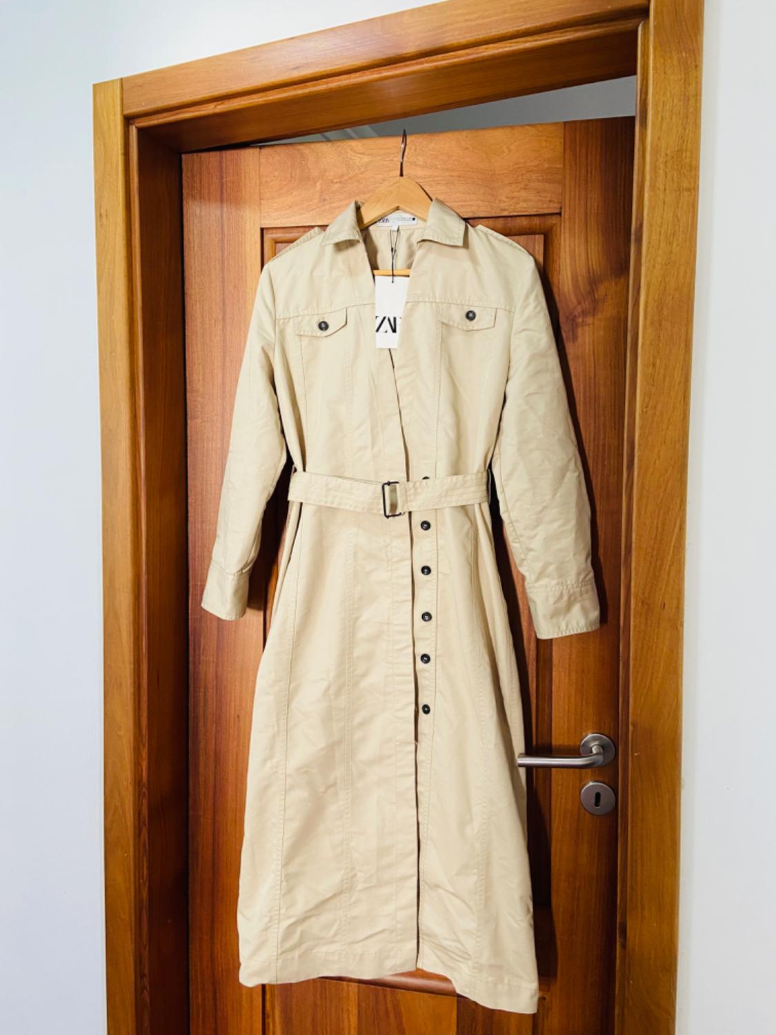 Front view of beige trench coat with belt and black button fastening.