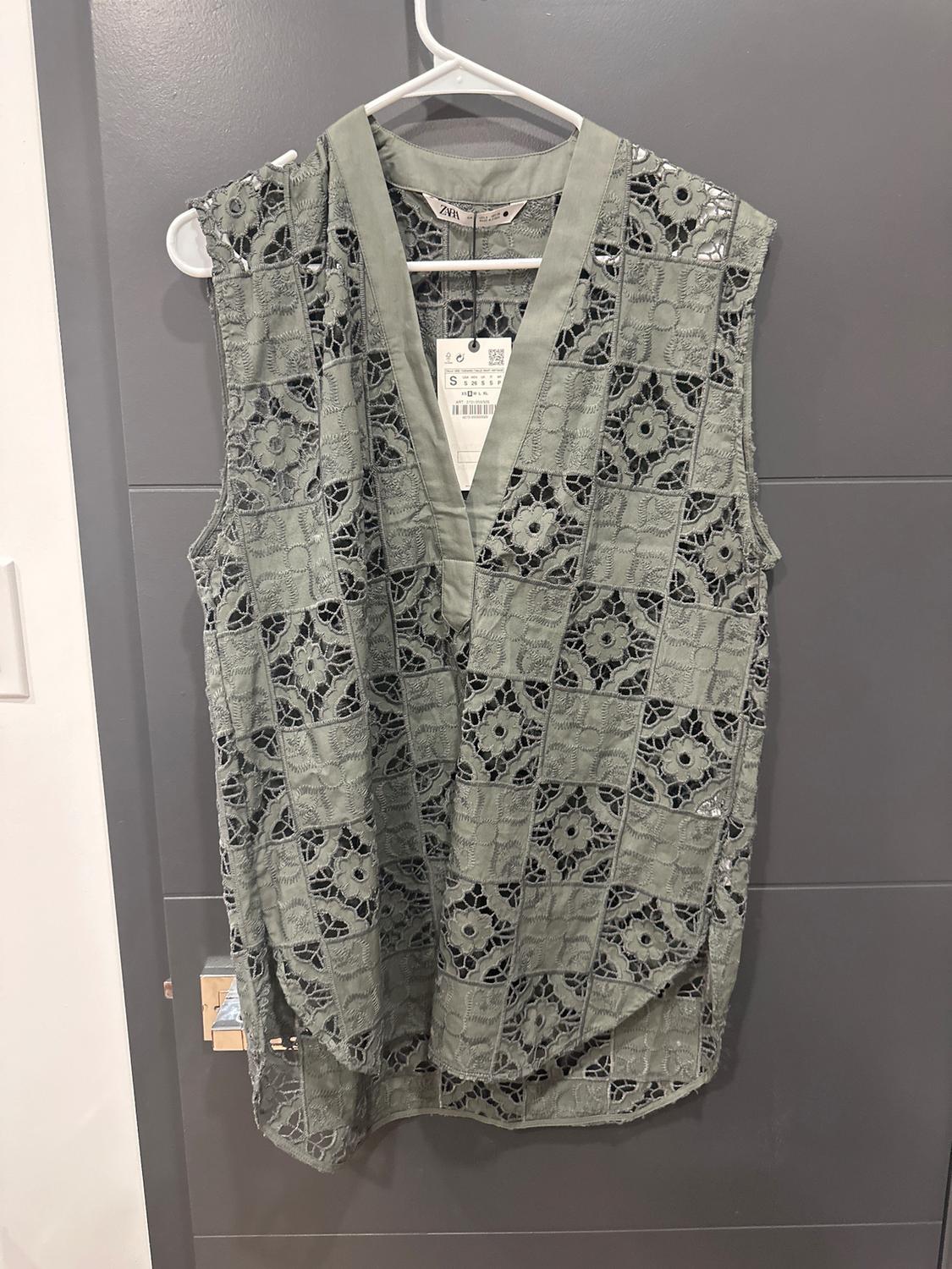 Green lace sleeveless blouse hanging on a white hanger against a gray wall. Visible tag with product details.