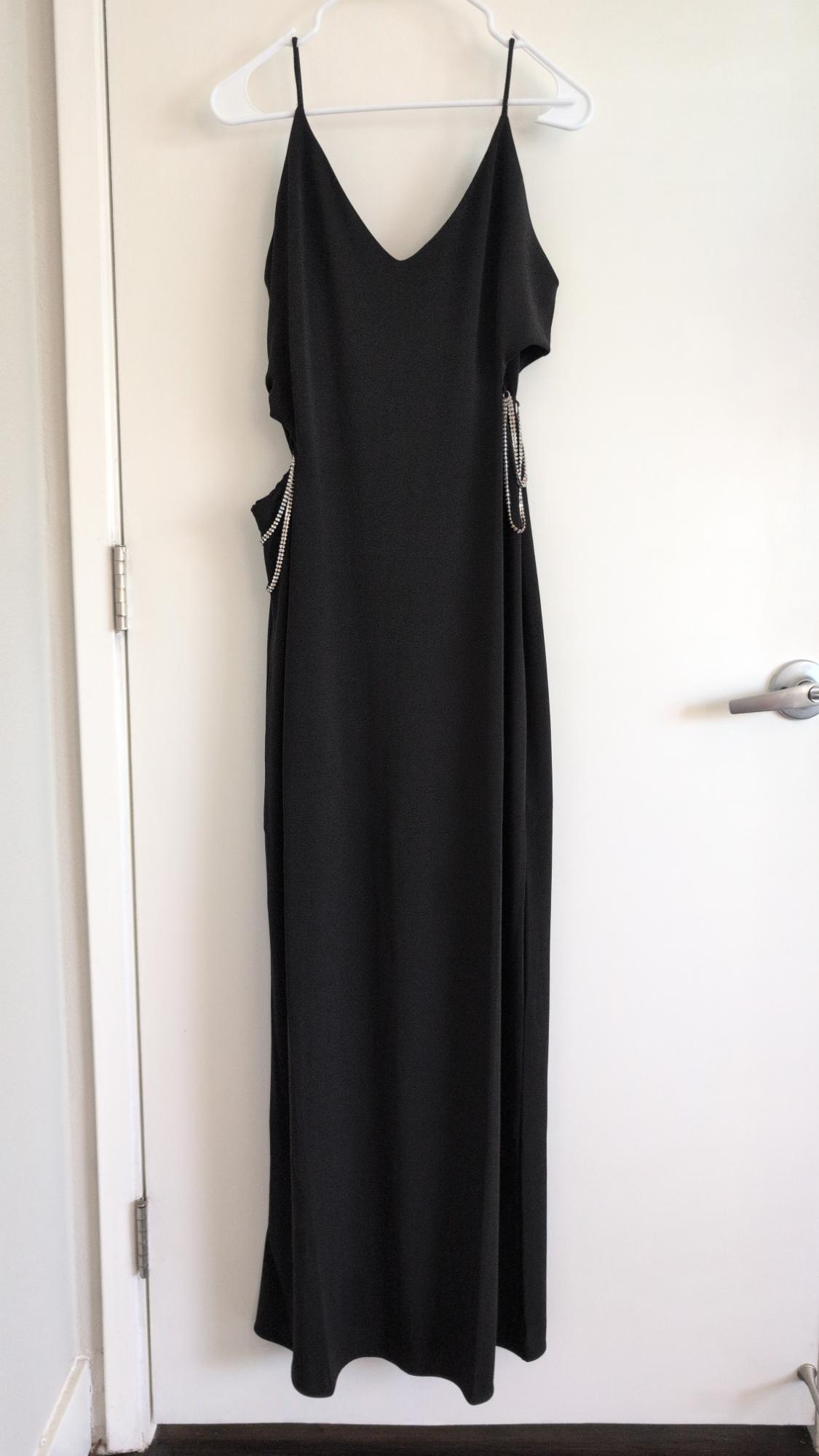 Black maxi dress with spaghetti straps, a V-neckline, and side slits with silver chains, hanging on a hanger on a white door.