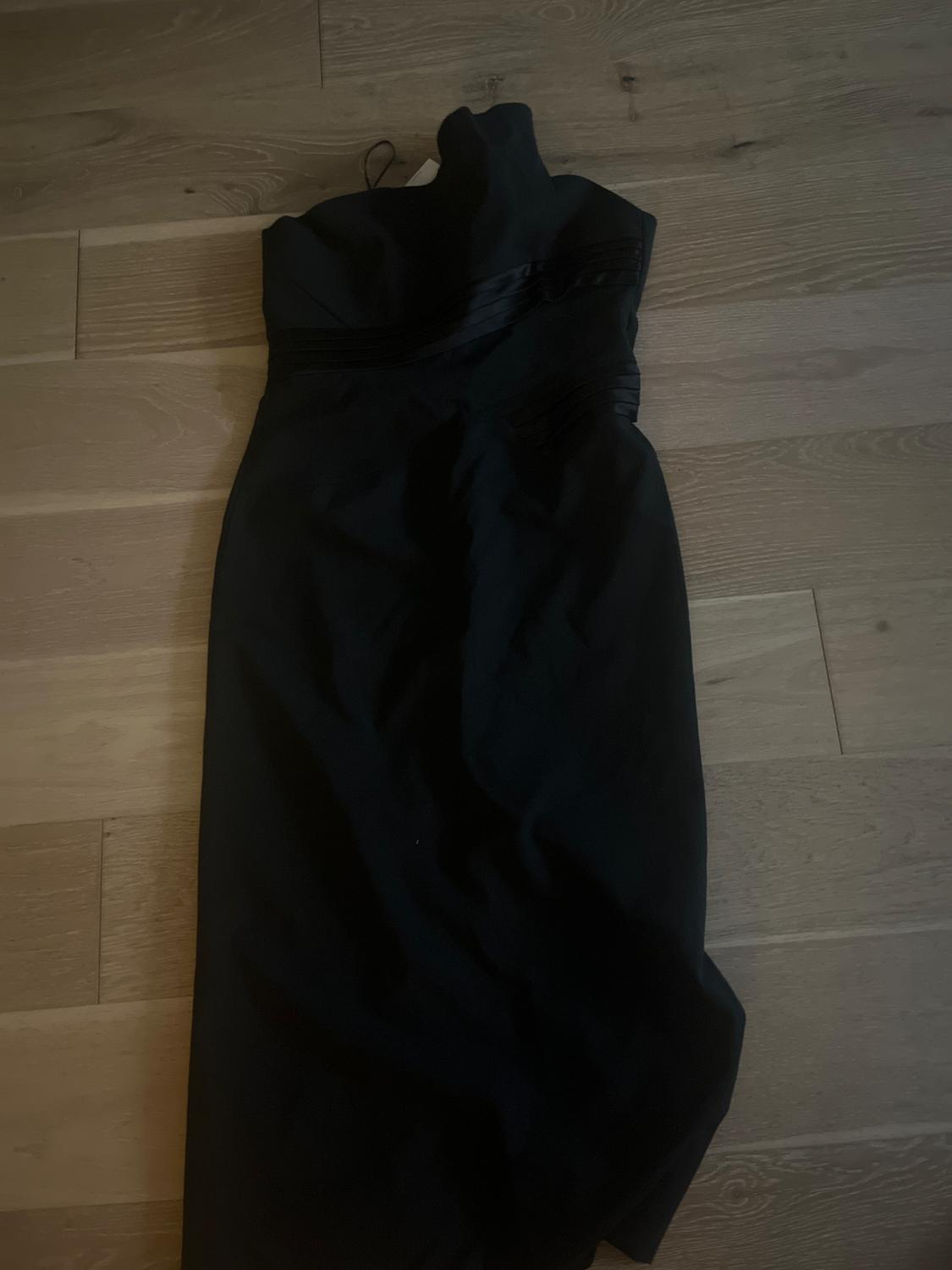 Sleeveless black dress with a high neck, ribbon detail at the waist, on a light wooden floor.