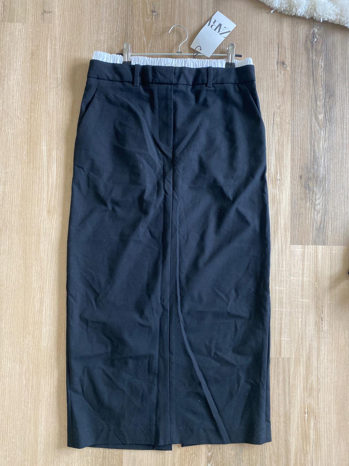 Long black skirt with a white elastic waistband, hanging on a hanger over a light wood floor. Tag visible at the top.
