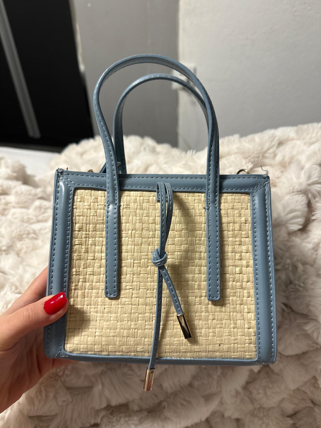 Small handbag with blue handles and a wicker body. White fabric background. Red painted nails holding the bag.