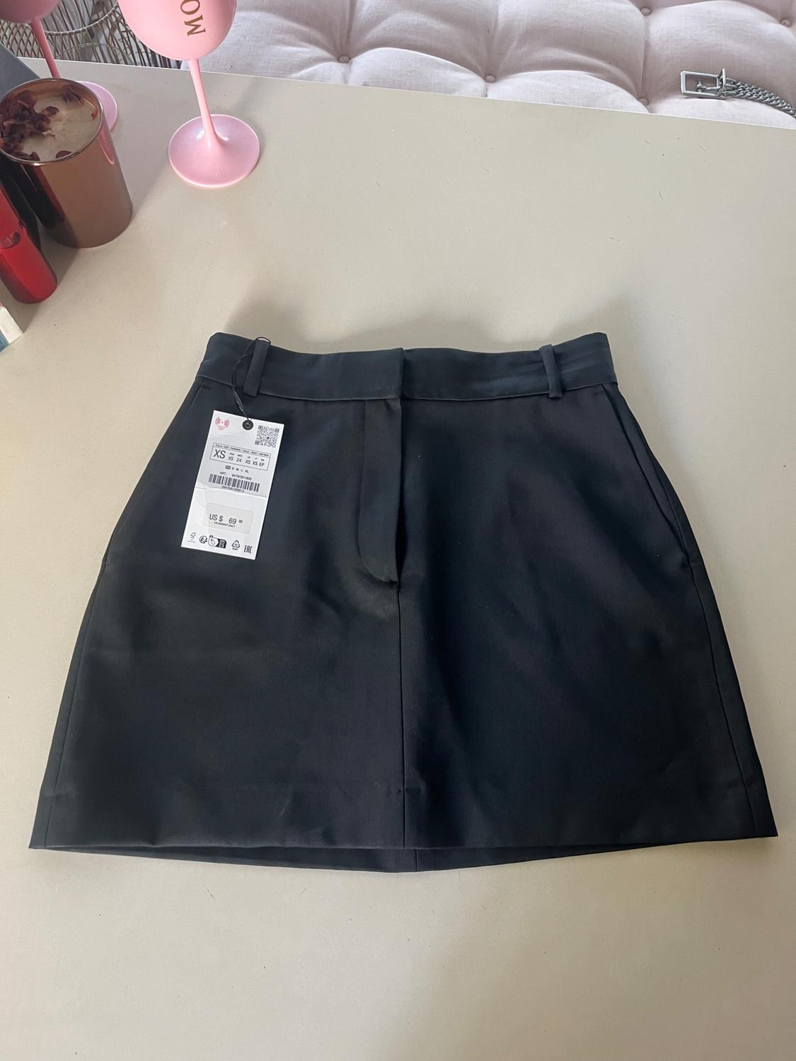 Black skirt on a beige table with an XS size tag and a price of $69. Background with pink glasses and a white quilted sofa.