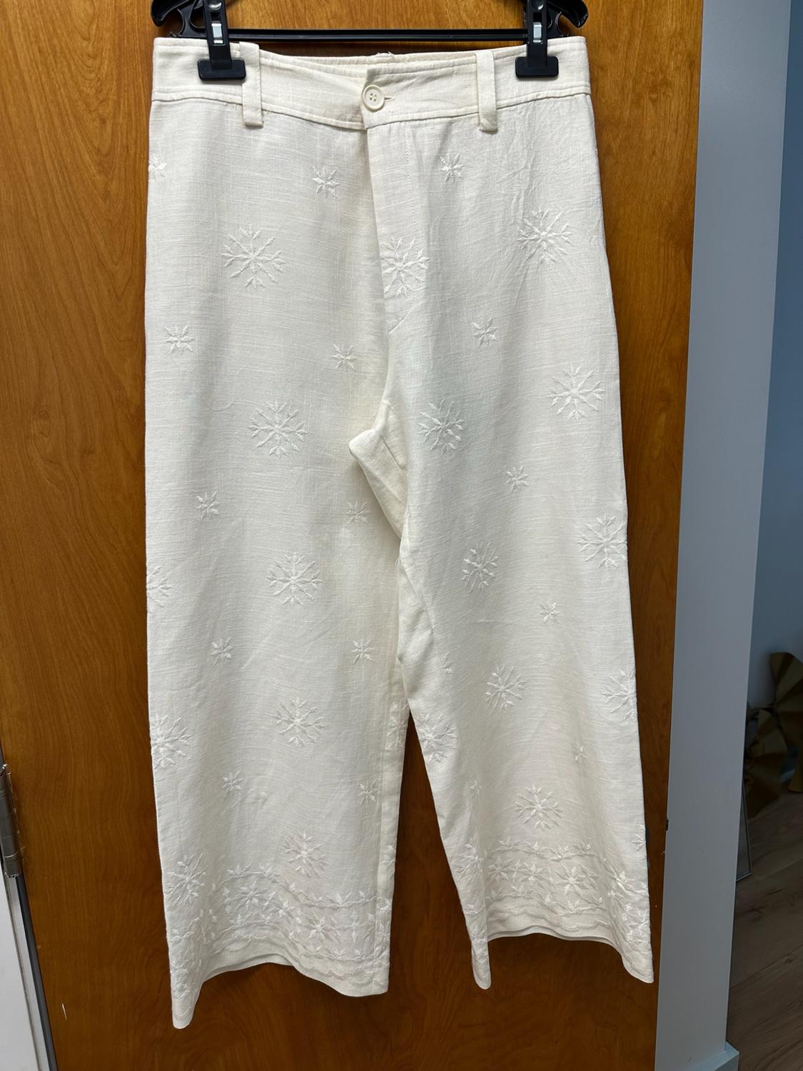 Cream wide-leg trousers with star embroidery and detailing on the hem, hanging on a black hanger over a wooden door.