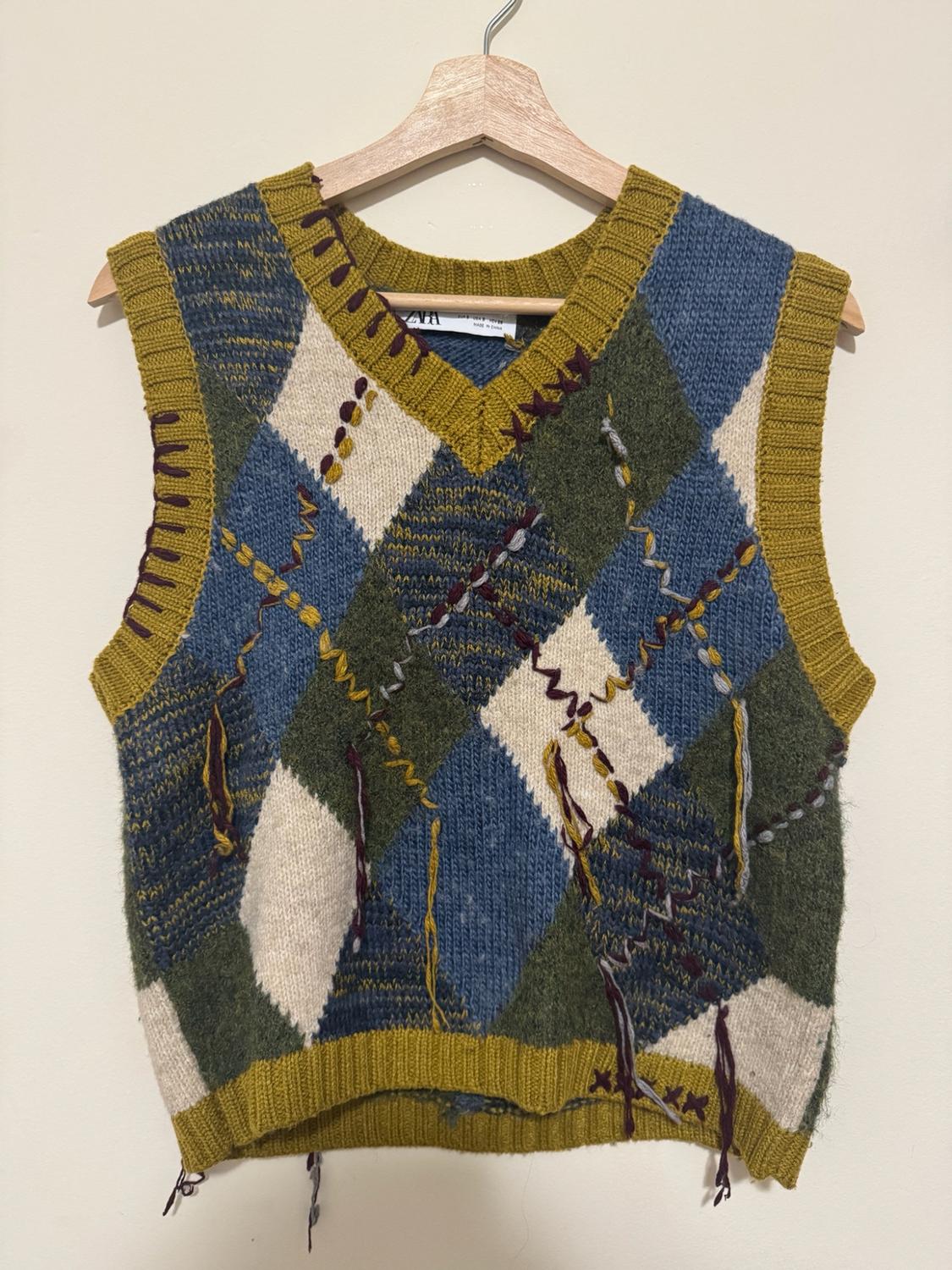 V-neck knit vest on a wooden hanger. Blue, green, and white argyle pattern, mustard trim, and loose threads. ZARA label.