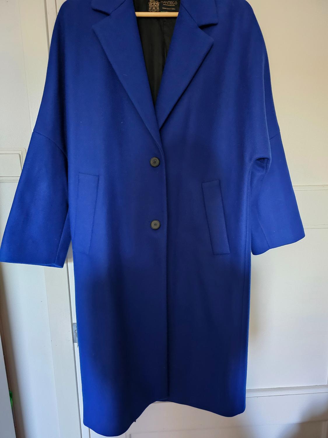 Long blue coat hanging on a hanger in front of a white door. Design with lapels, two buttons and pockets. Label bearing the text MANTECO.