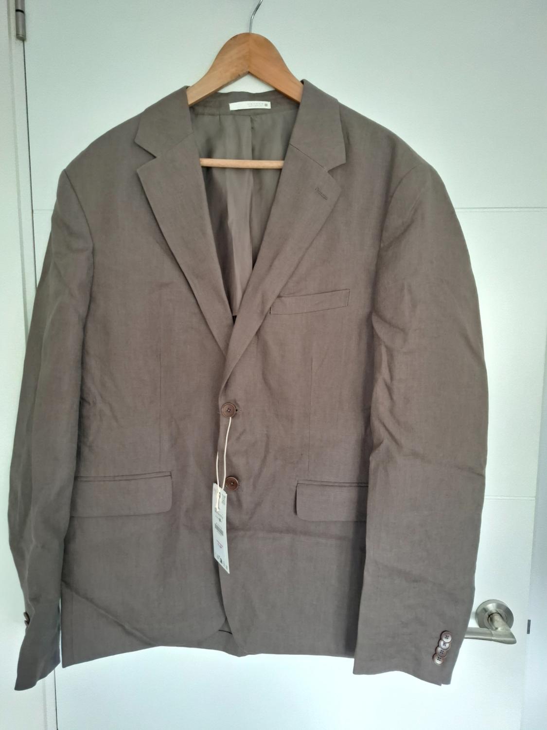 Brown linen jacket with classic lapels, two front buttons and pockets, hanging on a wooden hanger in front of a white door.