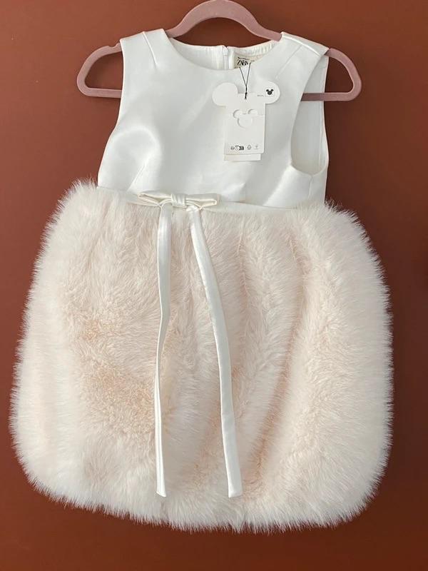 White children's dress with a beige fluffy skirt and bow, hanging on a pink hanger against a brown background. Mouse-shaped tag.