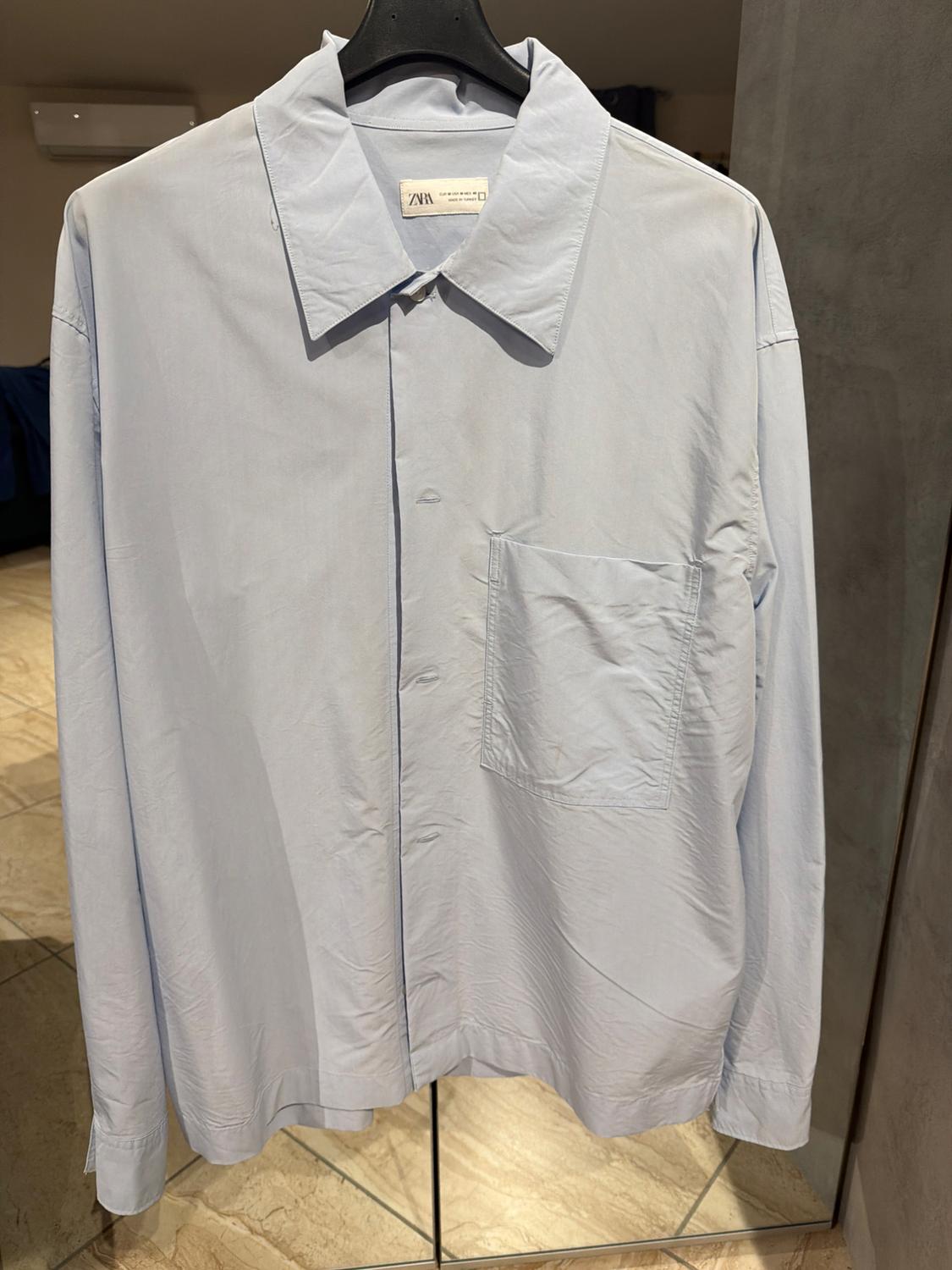 Light blue shirt hanging on a black hanger, with a front pocket and visible Zara label. Shop interior background.