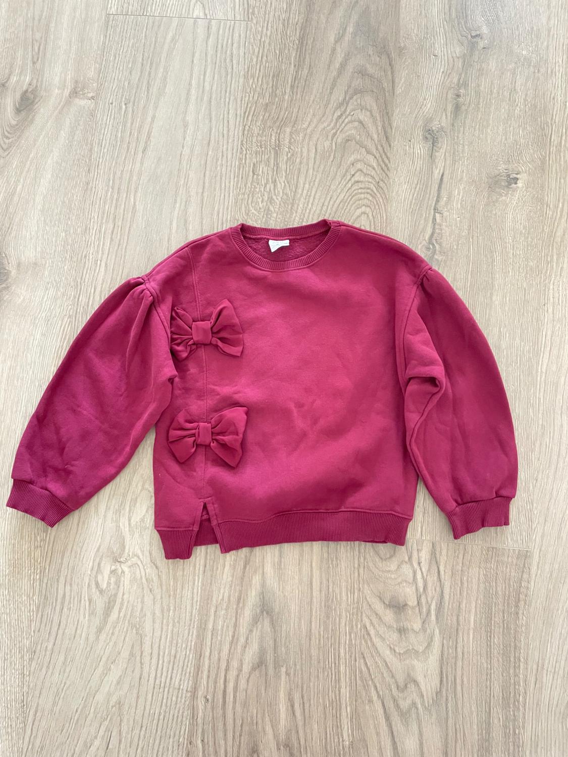 Pink sweatshirt with two bows on the left side, on a light wood floor. Long sleeves and round neck.